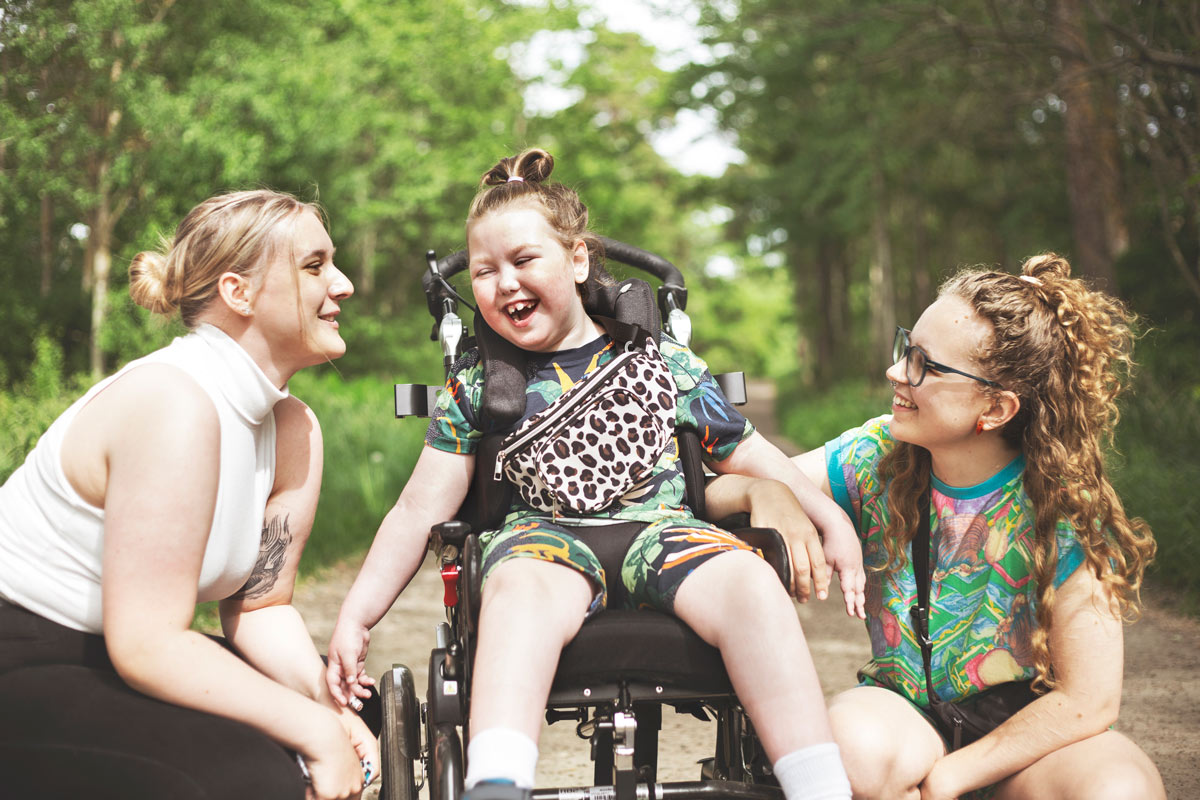 En flicka i rullstol skrattar tillsammans med två tjejer som jobbar som personliga assistenter.