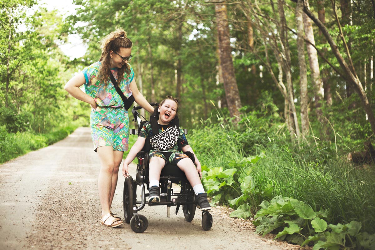 En skrattande flicka i rullstol är ute på promenad med sin personliga assistent. Det är en sommardag.